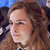Pixelated close-up of a person with light brown curly hair, neutral expression, and visible ear, against a blurred background with blue and dark tones.