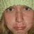 A static image of a person with light brown hair wearing a green knit hat, positioned against a blurred background.