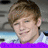 Pixelated close-up of a person with light brown hair, smiling, wearing a white shirt with a purple border at the bottom.