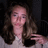 A pixelated image shows a person with long brown hair wearing a white top and dark jacket, making a peace sign gesture against a dark background.