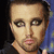 A person with dark hair and heavy eye makeup wears a black shirt against a blue background with yellow dots.