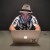 Pixelated figure wearing light-colored hat and patterned shirt, seated with arms resting on a rounded object against dark background.