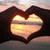 A heart-shaped frame of hands holds a sunset over water, with the sky shifting from pink to orange and the water reflecting golden light.