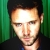 Pixelated close-up of a person with short dark hair against green vertical stripes, wearing dark clothing and blue collar.