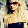 A static image of a person with long blonde hair wearing dark sunglasses and a black strapless top, set against a blurred background.