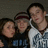 Three people pose together in a group photo with one person wearing a white shirt featuring blue text and another wearing a dark shirt with visible lettering.