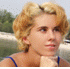 A person with short blonde hair in a blue strap top, shown from the shoulders up against an outdoor background.