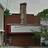 Static pixelated image of a building with a tall chimney, brown brick facade, and white trim, surrounded by greenery under a light sky.