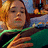 Pixelated static image of a person with light brown hair wearing a green and dark blue top, set against an indistinct background with warm and cool tones.