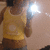 A person in a yellow sleeveless top and dark shorts stands against a light background with a bright light reflection on the left side.