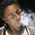 A person with short dark hair exhales white smoke while wearing a light-colored sleeveless shirt against a black background.