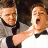 Pixelated static image of two men in formal attire with one man extending his arm toward the other's face.