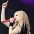 A woman with long blonde hair sings into a red microphone, raising her right arm against a dark background.