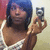 A person with blue hair holds a silver camera, wearing a white and brown patterned top against a red and beige background.