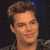 Pixelated close-up of a person with styled brown hair, light skin, and a faint smile against a dark background.