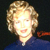 Pixelated image of a person with light brown curly hair wearing a dark blue top against a dark background with a red object on the right.