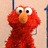 Red fuzzy character with large white eyes and orange nose stands against light background, surrounded by red pixelated text.