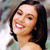 A pixelated close-up of a smiling person with dark hair, light skin, and visible teeth, wearing a light-colored top against a blurred background.