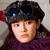 A pixelated close-up of a person wearing a dark gray flat cap with blue reflective spots, long dark hair, and a purple garment against a light gray background.