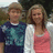 Two people stand side-by-side in a pixelated outdoor scene with greenery and a light-colored structure behind them.
