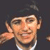 Pixelated image of a person with light skin tone wearing a dark jacket and white shirt, raising their right hand in a waving gesture against a dark background.