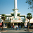 Pixelated static image of a building with a tall central spire, green roof sections, and palm trees under a clear blue sky.
