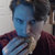 Pixelated static image of a person with dark hair, blue face, and maroon clothing against a light background.