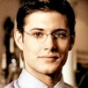 Static image of a person with dark hair, wearing round eyeglasses and a light-colored collared shirt, set against a blurred background with warm tones.