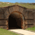 Pixelated image of a dark arched tunnel entrance with brown stone walls, set in a grassy landscape under a blue sky.