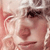 Pixelated close-up of a person's face with light skin tone, dark eyes, and a neutral expression, set against a blurred background with pink and white tones.