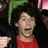 Pixelated image of a person with short dark hair wearing red clothing, mouth open in an expressive gesture against a dark background.