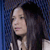 Pixelated close-up of a person with long dark hair against a blue patterned background, wearing a light-colored top.