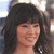 A static image of a person with long dark hair and bangs, smiling, against a blurred background with light and dark tones.