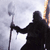 Silhouetted figure in dark clothing holding a long staff with a flat, rectangular top against a light gray background.