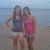 Two women stand on a beach with water and sky in the background; one wears a dark sleeveless top and light shorts, the other a pink sleeveless top and light shorts.