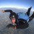 Pixelated figure in dark wetsuit with blue accents swimming underwater, arms extended forward and legs trailing behind against gradient sky-blue background.
