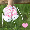 A pair of pink and white sneakers with white laces hangs from a wooden surface above green grass, with a pink heart symbol in the lower right.