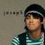 Pixelated image of a person wearing dark cap with white stripe, green and white checkered shirt, and dark vest against muted background.