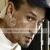 Pixelated close-up of a person's face with visible ear, dark hair, and light skin tone, wearing dark clothing against a blurred background.
