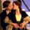Two figures in close embrace, one with dark hair and black top, the other with light brown hair and yellow-accented black clothing against a blurred background.