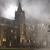 Pixelated cityscape with tall building and spire under overcast sky, featuring brown and gray tones with scattered light-colored windows.