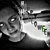 Pixelated grayscale image of a stylized character with a large head, wide eyes, and a broad smile, holding a green object against a dark background.