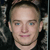 Pixelated close-up of a person's face with light skin, blue eyes, and short brown hair against a dark background.