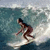 A pixelated figure surfs on a wave, body angled left with arms extended, wearing dark hair and a brown swimsuit against blue-green water and white foam.