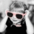 Pixelated static image of a person wearing dark sunglasses with light pink frames, black top, and gray hair against a gradient background.