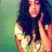 Pixelated image of a person with long dark hair wearing a light-colored top, making a peace sign gesture against a green gradient background.