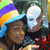 Two people in a close-up pose with one wearing a colorful striped hat and the other in a white mask, set against a blurred background.