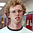 Pixelated image of a person with curly light brown hair wearing a white shirt with black collar, set against blurred background with purple text overlays.