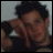Pixelated static image of a person with dark hair and brown skin tone, cropped to show upper body against light background.
