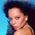 Pixelated close-up of a person with dark curly hair against a blue background, wearing a light-colored earring and a thin dark necklace.